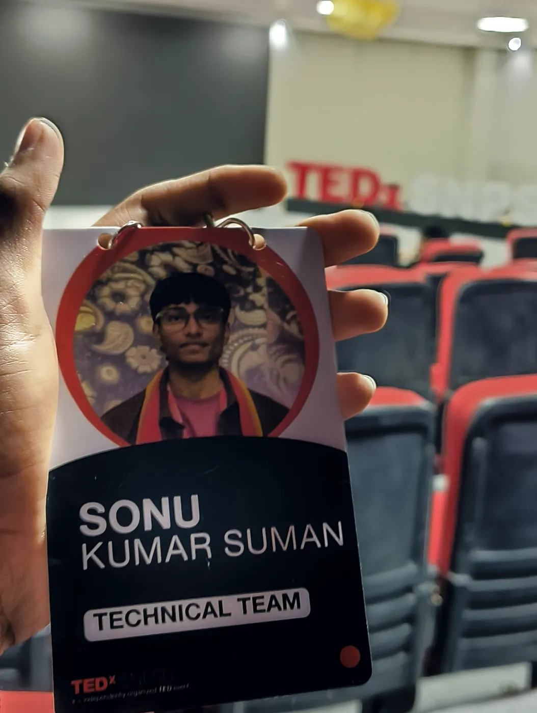 TEDx technical team badge with the name Sonu Kumar Suman, held up in an auditorium with red seats