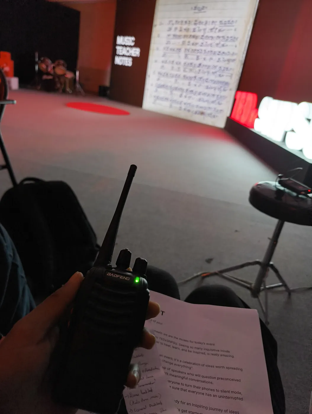 Walkie-talkie and a printed run sheet held backstage, with the red-lit TEDx stage in the background