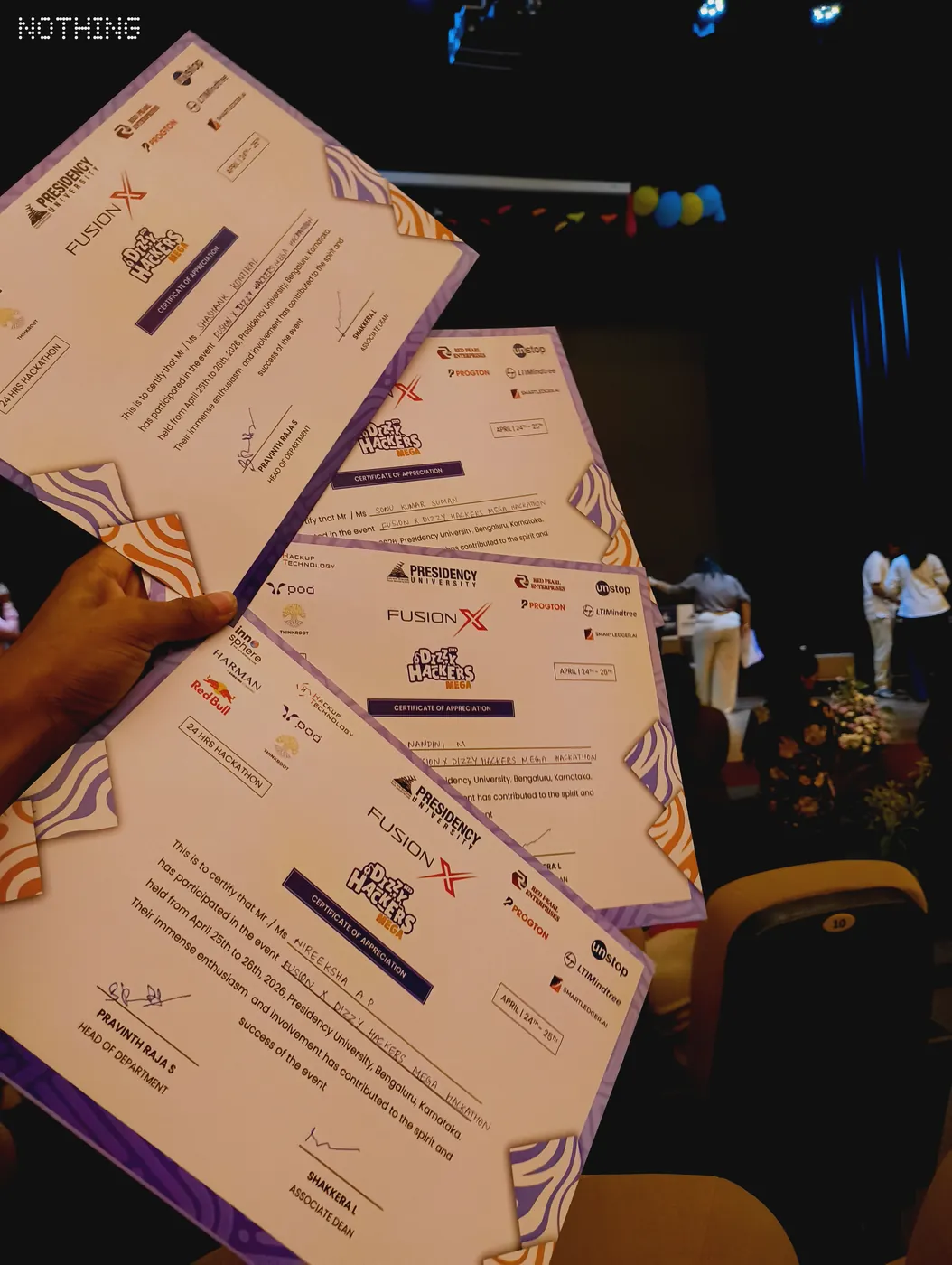 Four participation certificates fanned out in hand inside an auditorium