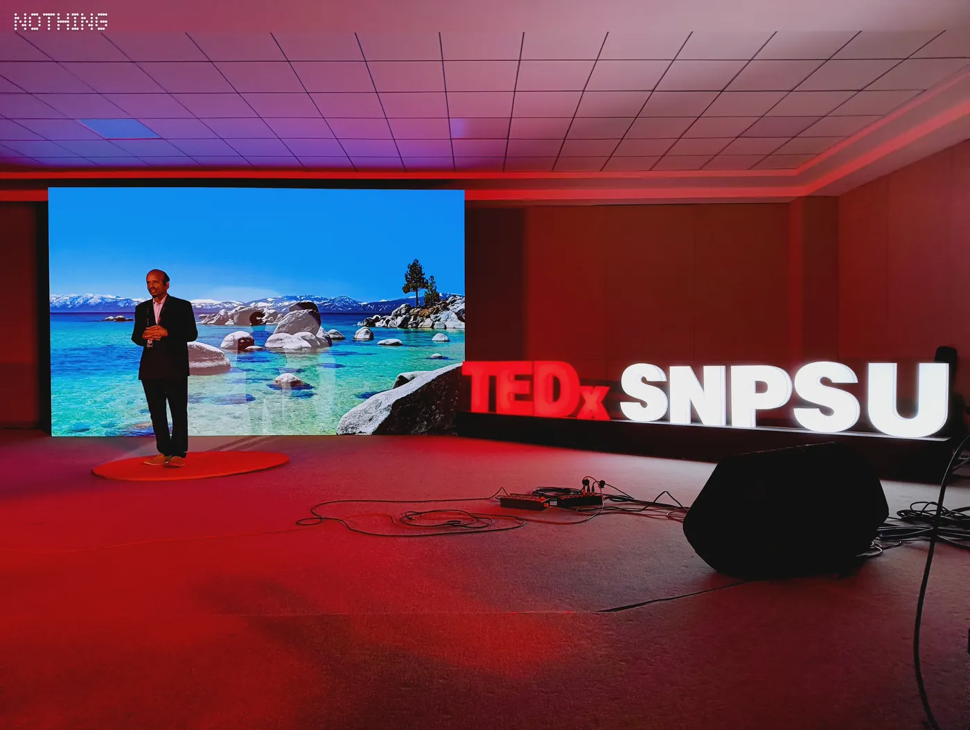 Speaker on the red-lit TEDxSNPSU stage beside the large TEDxSNPSU letters